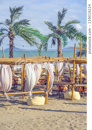 Beach lounge area with bamboo cabanas, curtains on sandy shore with palm trees and sea horizon. Beach resort relaxation with lounge seating, tropical vacation destination 139019224