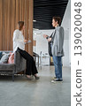 Two businesswomen greeting and talking in front of gray sofa in open modern office. Greeting, discussion, office, teamwork, business. 139020048