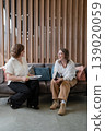 Women in professional attire conversing and discussing on sofa in modern business lounge. Conversation, discussion, lounge, women, professional, business. 139020059