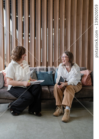 Women in professional attire conversing and discussing on sofa in modern business lounge. Conversation, discussion, lounge, women, professional, business. Women in professional attire conversing and discussing on sofa in modern business lounge. Conversation, discussion, lounge, women, professional, business. 139020059