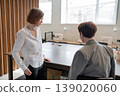 Team members consulting one another by open communal area in modern workplace with wood detail. Consulting, communal, modern, workplace, teamwork, women. 139020060