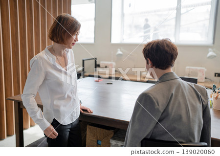 Team members consulting one another by open communal area in modern workplace with wood detail. Consulting, communal, modern, workplace, teamwork, women. Team members consulting one another by open communal area in modern workplace with wood detail. Consulting, communal, modern, workplace, teamwork, women. 139020060