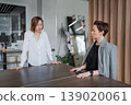 Female team members standing and talking at office desk in bright collaborative workspace. Team, talking, desk, office, collaborative, businesswomen. 139020061