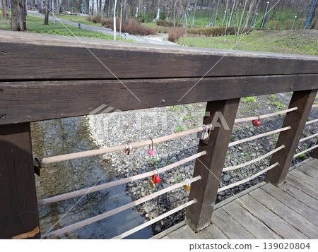 Locks are attached to the rope railing of a wooden bridge. The bridge is over a stream in a park with trees and grass visible. People pass by the area to enjoy the scenery. 139020804