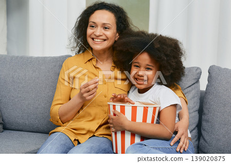 African American mother with her little daughter hugging and watching cinema together and eating popcorn from a large striped bucket 139020875