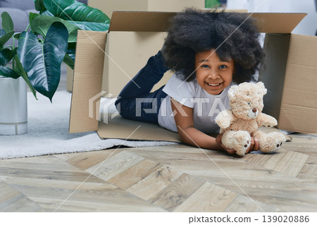 Happy African American little girl playing with cardboard boxes and her plush bear, having fun while moving new home Happy African American little girl playing with cardboard boxes and her plush bear, having fun while moving new home 139020886