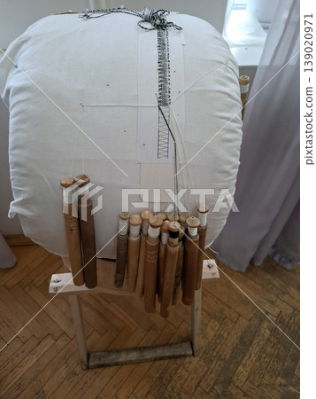 A lace maker sits at a lace pillow with wooden bobbins and thread, creating detailed lace patterns. The workspace shows tools and techniques of the craft. The setting is bright and organized. 139020971