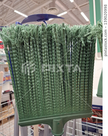 The thick, textured green bristles of a plastic garden broom are displayed on a shelf in a retail store. Bright lighting shows the details of the bristles. The thick, textured green bristles of a plastic garden broom are displayed on a shelf in a retail store. Bright lighting shows the details of the bristles. 139020998