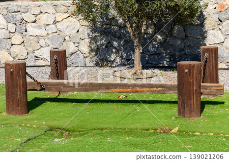 A rustic balancing beam made of wood and chains in a sunlit park surrounded by stone walls and grass A rustic balancing beam made of wood and chains in a sunlit park surrounded by stone walls and grass 139021206