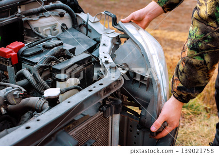 Worker works on car by taking off old headlight assembly near engine in garage setting. 139021758