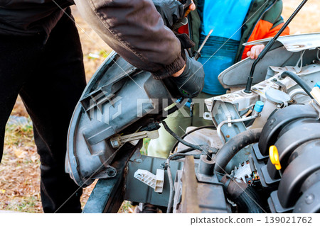 Service worker removes headlight part from car near engine while process outdoors Service worker removes headlight part from car near engine while process outdoors 139021762