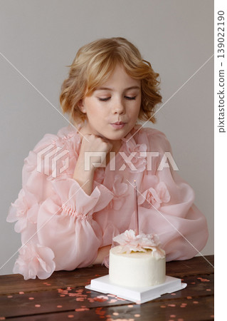 Smiling girl celebrating birthday with cake and candle on wooden table, white wall Smiling girl celebrating birthday with cake and candle on wooden table, white wall 139022190