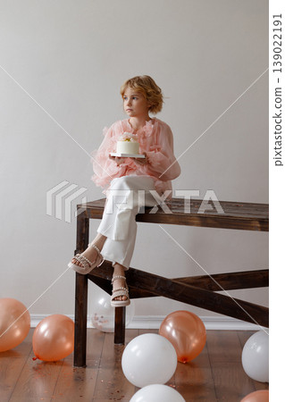 Smiling girl holding birthday cake sitting on wooden bench in room with white wall 139022191