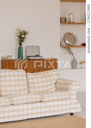 Beige plaid sofa standing in living room with white wall, wooden cabinet and flowers Beige plaid sofa standing in living room with white wall, wooden cabinet and flowers 139022460