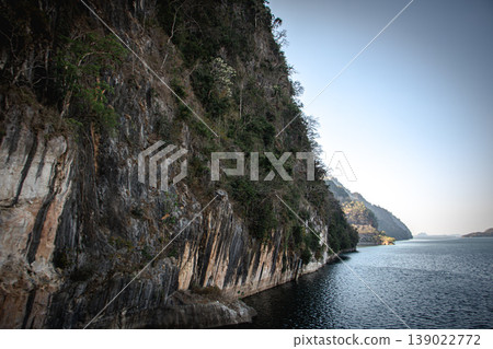 Dramatic rock cliff along calm mountain lake under clear sky, scenic wilderness landscape with textured stone wall and copy space. Dramatic rock cliff along calm mountain lake under clear sky, scenic wilderness landscape with textured stone wall and copy space. 139022772
