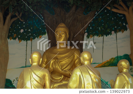 Golden Buddha statue teaching disciples under Bodhi tree mural inside public Thai temple, spiritual Buddhist art and culture. Golden Buddha statue teaching disciples under Bodhi tree mural inside public Thai temple, spiritual Buddhist art and culture. 139022973