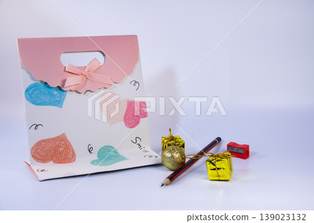 Decorative gift bag with heart patterns, pencil, sharpener and small golden gift boxes isolated on white background. Still life concept for celebration, love, stationery and education. Decorative gift bag with heart patterns, pencil, sharpener and small golden gift boxes isolated on white background. Still life concept for celebration, love, stationery and education. 139023132