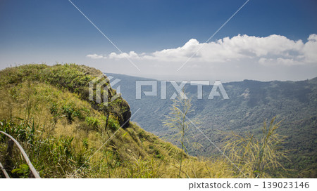 Grassy mountain cliff overlooking vast green valley under blue sky. Scenic highland landscape with wild vegetation, symbolizing adventure, hiking, exploration and untouched nature. Grassy mountain cliff overlooking vast green valley under blue sky. Scenic highland landscape with wild vegetation, symbolizing adventure, hiking, exploration and untouched nature. 139023146