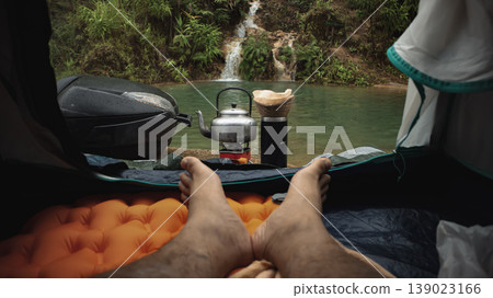 POV from inside camping tent with feet resting on sleeping pad, coffee kettle and dripper by river waterfall. Peaceful morning outdoor lifestyle, adventure travel and nature relaxation. POV from inside camping tent with feet resting on sleeping pad, coffee kettle and dripper by river waterfall. Peaceful morning outdoor lifestyle, adventure travel and nature relaxation. 139023166