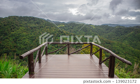 Wooden viewpoint deck overlooking lush green mountain forest under cloudy sky. Scenic tropical highland landscape, peaceful nature travel destination and outdoor adventure scenery. Wooden viewpoint deck overlooking lush green mountain forest under cloudy sky. Scenic tropical highland landscape, peaceful nature travel destination and outdoor adventure scenery. 139023172