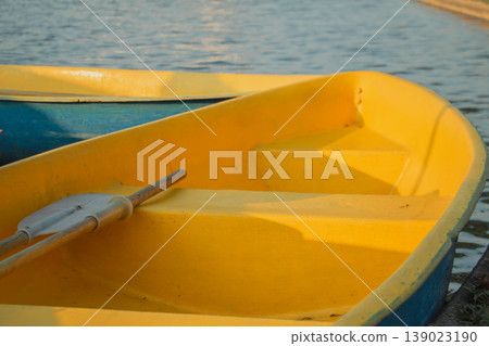 Yellow rowboat with oar resting by calm water under warm sunlight. Minimal outdoor scene with copy space, ideal for travel, summer leisure, relaxation and waterfront themes. Yellow rowboat with oar resting by calm water under warm sunlight. Minimal outdoor scene with copy space, ideal for travel, summer leisure, relaxation and waterfront themes. 139023190