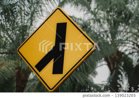 Yellow diamond traffic sign showing side road merging from left. Road warning symbol against palm tree background, transportation and driving safety concept outdoors. Yellow diamond traffic sign showing side road merging from left. Road warning symbol against palm tree background, transportation and driving safety concept outdoors. 139023203