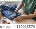Hands cutting thread from old jeans for reuse, promoting sustainable fashion and circular economy principles. Person upcycling denim jeans with scissors, repairing clothing 139023579