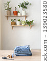 Stack of old blue jeans ready for upcycling on a wooden table with scissors and thread. Potted plants decorating shelves in background. Reusing things. Environmental awareness 139023580