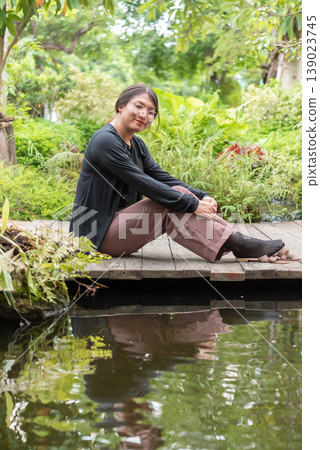 LGBTQ woman sitting by pond in lush garden wearing glasses relaxed pose natural light peaceful outdoor setting reflecting calm identity and inclusive lifestyle LGBTQ woman sitting by pond in lush garden wearing glasses relaxed pose natural light peaceful outdoor setting reflecting calm identity and inclusive lifestyle 139023745