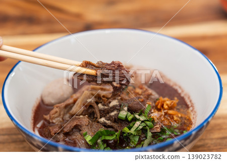 Thai boat noodle soup with beef slices meatball herbs and dark broth held by chopsticks in bowl on wooden table showing rich flavor and traditional street food style Thai boat noodle soup with beef slices meatball herbs and dark broth held by chopsticks in bowl on wooden table showing rich flavor and traditional street food style 139023782