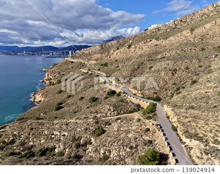 Meandering Coastal Route Hugging Jagged Cliffs Beneath Bright Sunlit Sky Over Deep Blue Ocean 139024914