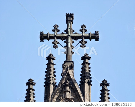 Decorative Church Pinnacle. Detailed Ornate Cross Mounted On Aged Gothic Tower Under Bright Blue Sky Decorative Church Pinnacle. Detailed Ornate Cross Mounted On Aged Gothic Tower Under Bright Blue Sky 139025153