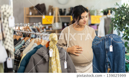 Pregnant Latin girl is busy looking at clothes items in store, she chooses jeans Pregnant Latin girl is busy looking at clothes items in store, she chooses jeans 139025999