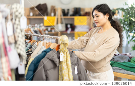 Happy pregnant latina female customer chooses coat or jacket in clothing market Happy pregnant latina female customer chooses coat or jacket in clothing market 139026014
