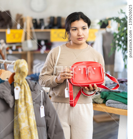 Positive latina female standing against background of shelves with clothes in modern store and choosing bag Positive latina female standing against background of shelves with clothes in modern store and choosing bag 139026025