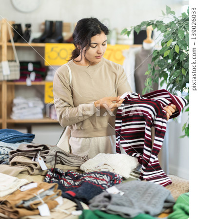 Latina female buyer standing in clothing market and carefully examines selection of sweaters on sale of winter clothing collection Latina female buyer standing in clothing market and carefully examines selection of sweaters on sale of winter clothing collection 139026033