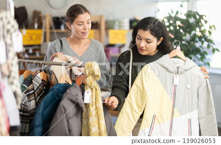 Two happy young women choosing coat or jacket for cold season in clothing shop with large assortment Two happy young women choosing coat or jacket for cold season in clothing shop with large assortment 139026053