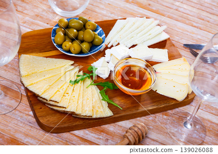 On wooden table there is cutting board with assorted cheese, honey, pickled olives - 139026088