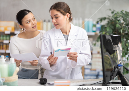 Portrait of young woman pharmacist helping asian female client with paper in hand looking for something or choosing Portrait of young woman pharmacist helping asian female client with paper in hand looking for something or choosing 139026210