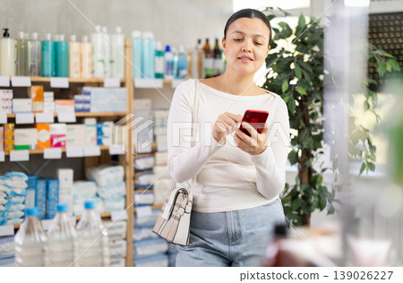 Middle-aged woman scrolling phone while looking for something in drugstore Middle-aged woman scrolling phone while looking for something in drugstore 139026227