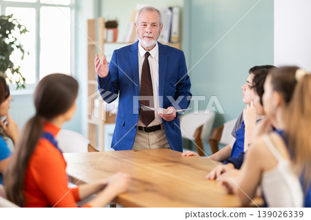 Mature male teacher addresses round table young workers meeting participants. Mature male teacher addresses round table young workers meeting participants. 139026339