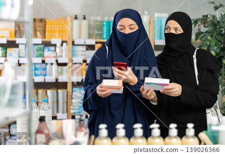 Attentive two woman in hijab choosing box with pills to improve well-being in pharmacy, scan QR code on label 139026366