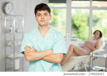 Young man doctor posing in doctor's cabinet with patient lying on clinical chair 139026421