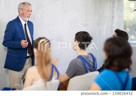 Mature male lecturer stands in front of young worker audience, delivers lecture Mature male lecturer stands in front of young worker audience, delivers lecture 139026432