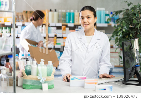 Portrait of young asian woman pharmacist standing at prescription counter and working on computer, stocktaking medicines at pharmacy shop and female assistant in background Portrait of young asian woman pharmacist standing at prescription counter and working on computer, stocktaking medicines at pharmacy shop and female assistant in background 139026475