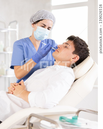 Closeup face of young male client receiving injections during lip enhancement procedure, professional cosmetologist hands in gloves holding syringe 139026516