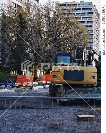 Street construction with a yellow excavator and orange safety barriers. Gravel and dirt cover the ground Street construction with a yellow excavator and orange safety barriers. Gravel and dirt cover the ground 139026673