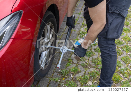 Man loosening wheel nuts with cross wrench during car tire change on pavement Man loosening wheel nuts with cross wrench during car tire change on pavement 139027357