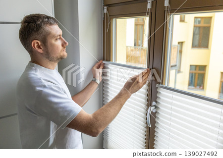 Man adjusting window blinds in modern apartment interior with soft natural light Man adjusting window blinds in modern apartment interior with soft natural light 139027492