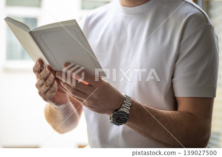 Close up of hands holding book while reading in soft daylight near window Close up of hands holding book while reading in soft daylight near window 139027500
